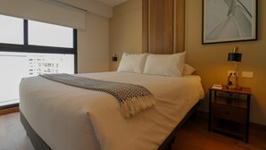 Et hotelværelse med en stor seng, et natbord med en lampe, et billede i ramme på væggen og et vindue med udsigt til bygninger.