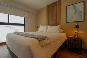 Quarto de hotel com uma cama grande, uma mesa de cabeceira com abajur, um quadro na parede e uma janela com vista para prédios.