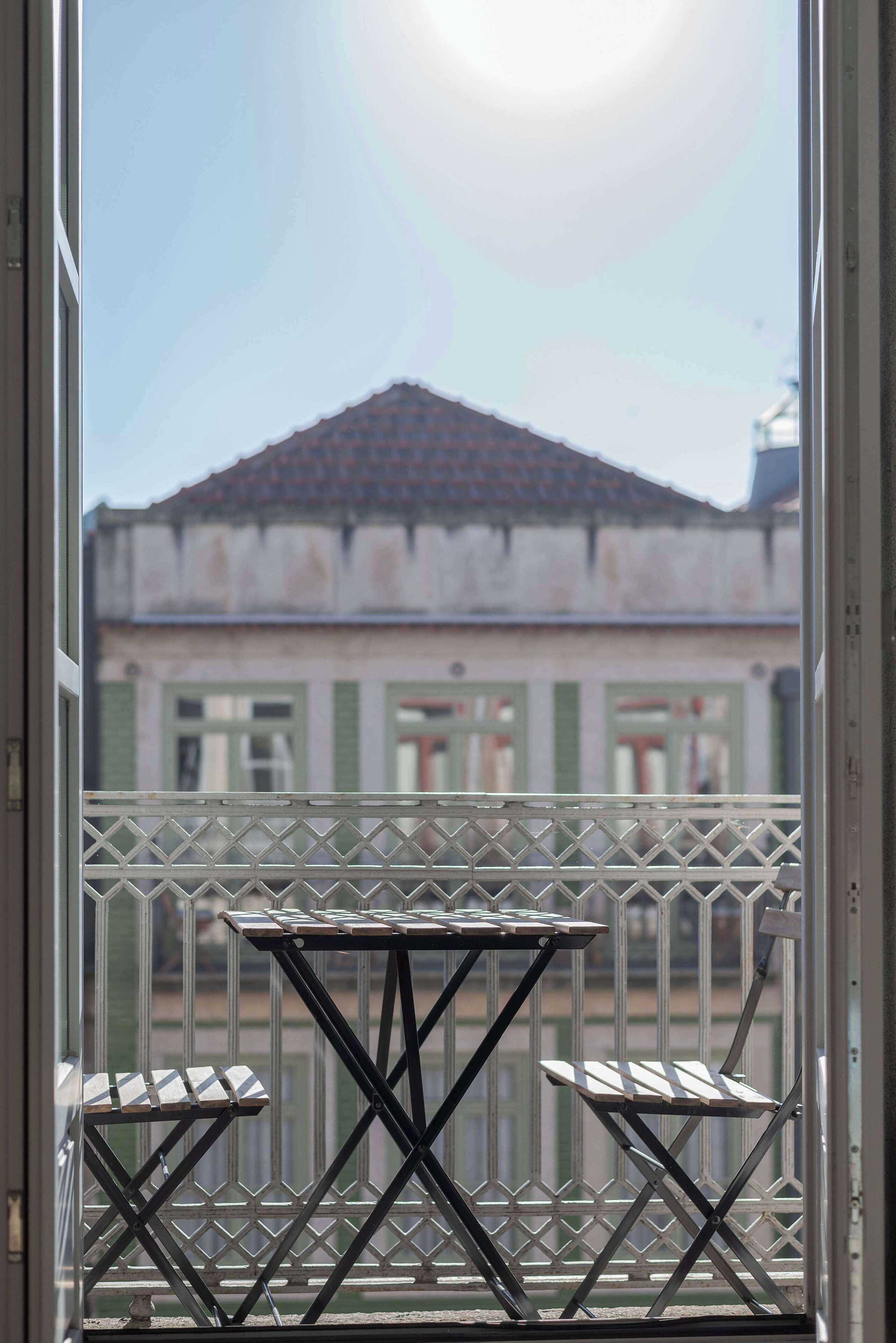 A balcony with a table and two chairs, overlooking a building with green shutters and a tiled roof.
