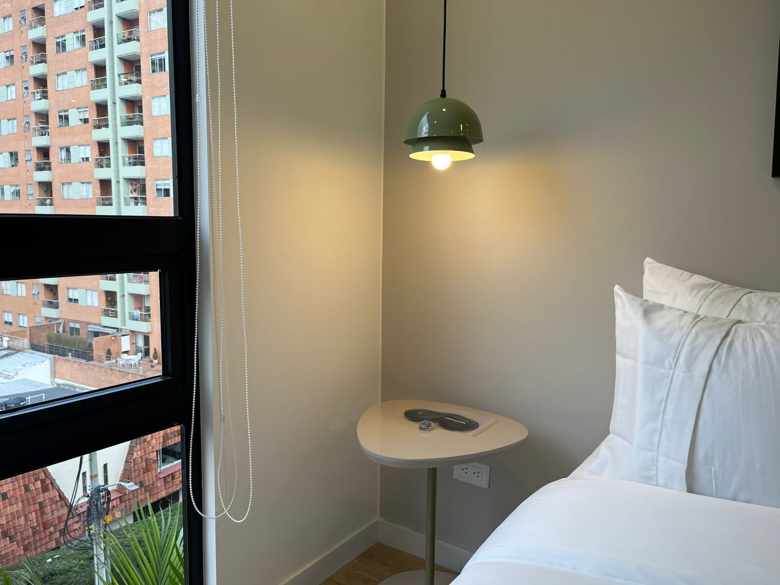 Une chambre d’hôtel avec un lit, une petite table ronde dotée d’une prise électrique, un luminaire suspendu vert et une vue sur un bâtiment en briques, visible par la fenêtre.