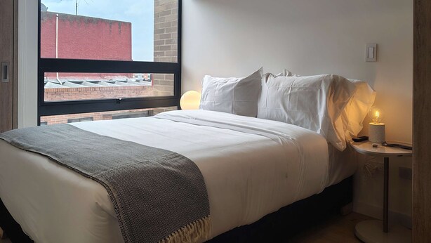A modern bedroom with a large window, a bed with white linens, a bedside table with a lamp, and a view of a brick building outside. - w Wonderful Loft in the Heart of Bogot (Bogotá)