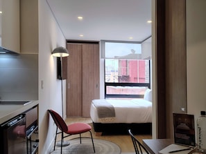 Ein modernes Hotelzimmer mit Bett, Schreibtisch mit Stuhl, einer kleinen Küchenzeile, einem großen Fenster mit Stadtblick und einer Deckenleuchte.