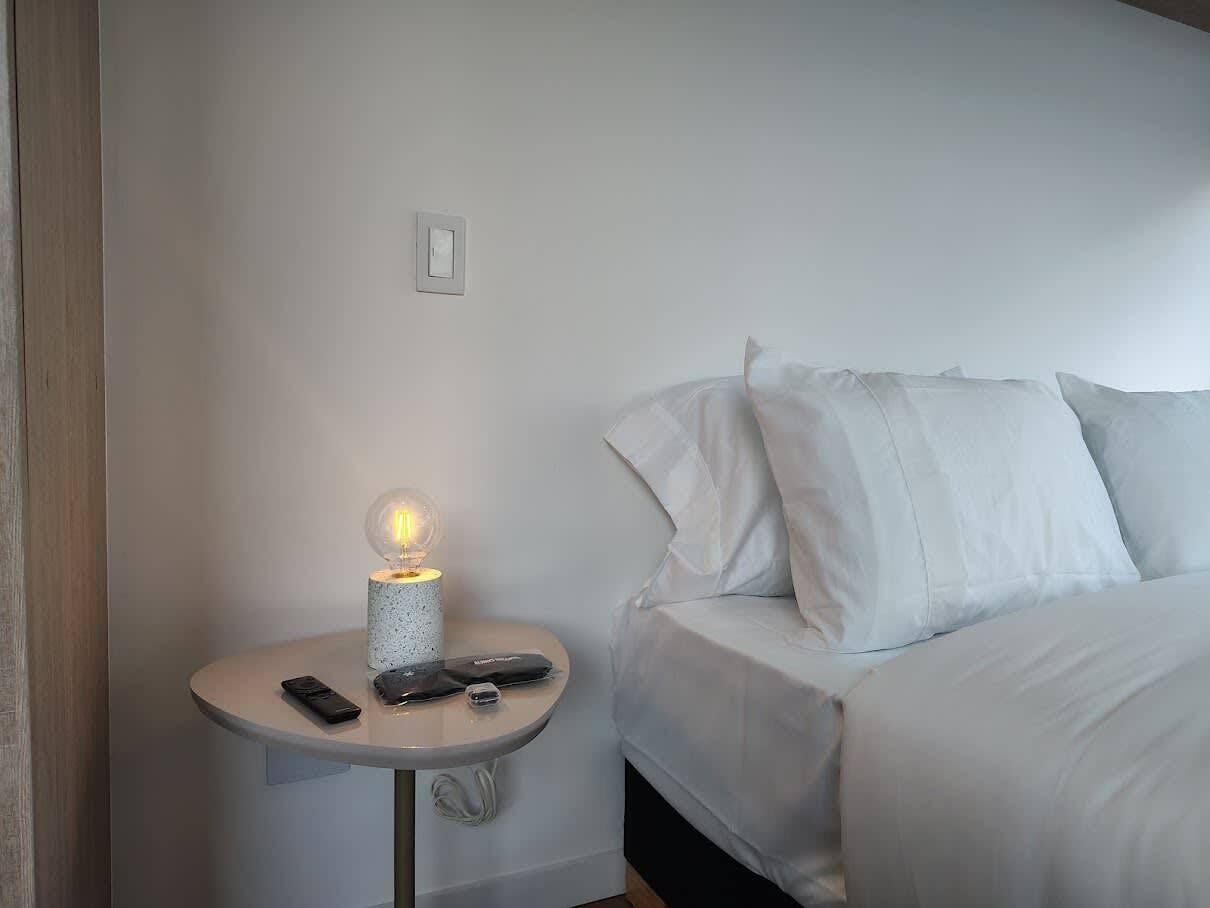 Ein ordentlich bezogenes Bett mit weißen Leinen, ein kleiner Nachttisch mit einer Lampe und einer Fernbedienung darauf.