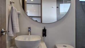 Et moderne badeværelse med en stor, oval håndvask, et rundt spejl, et håndklædestativ, en håndtørrer og et toilet.