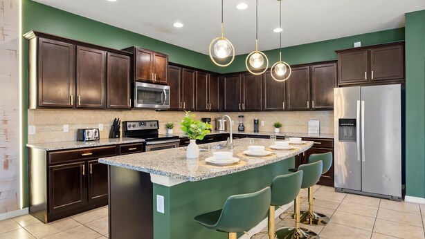 A modern kitchen with dark wood cabinets, stainless steel appliances, a granite countertop, and a central island with bar stools. - Upstay Epic Windsor Home w Pool Cinema Games (Kissimmee)