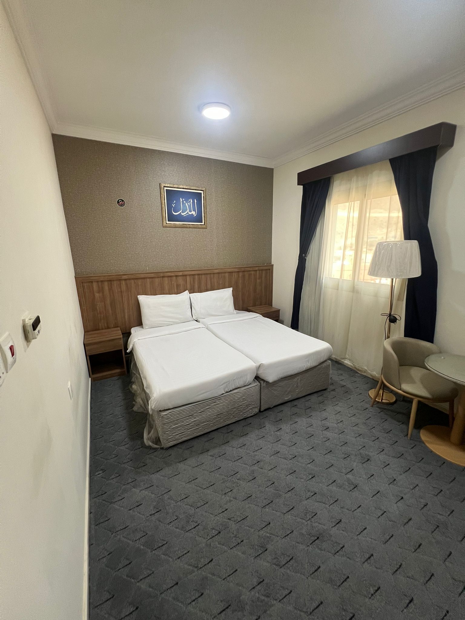 Quarto de hotel com cama, uma mesinha, uma cadeira, abajur e um letreiro fixado na parede.