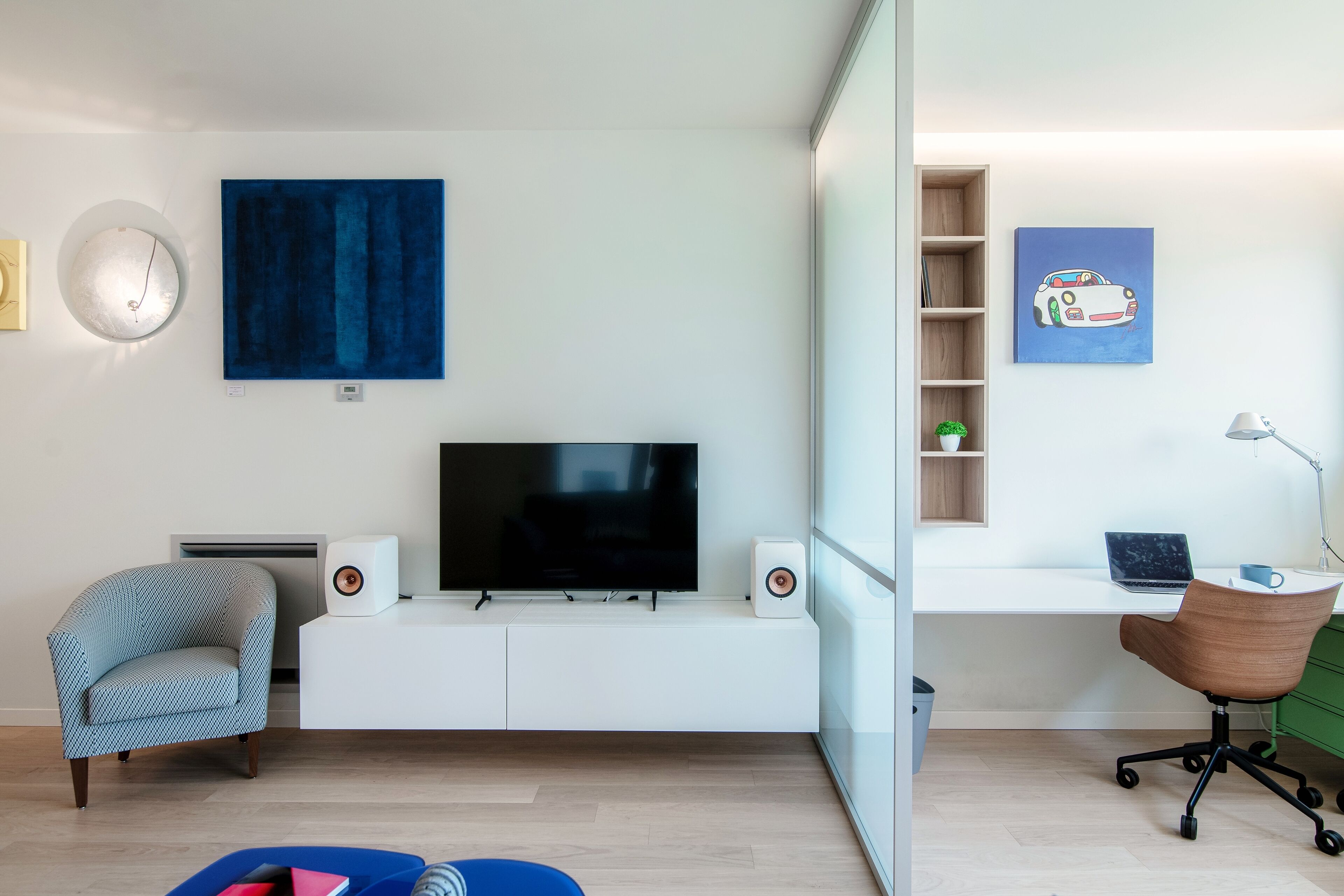 Una sala de estar moderna con un televisor de pantalla plana, una pared azul de acento, una silla de madera, un escritorio con una computadora portátil y un mueble de almacenamiento verde.