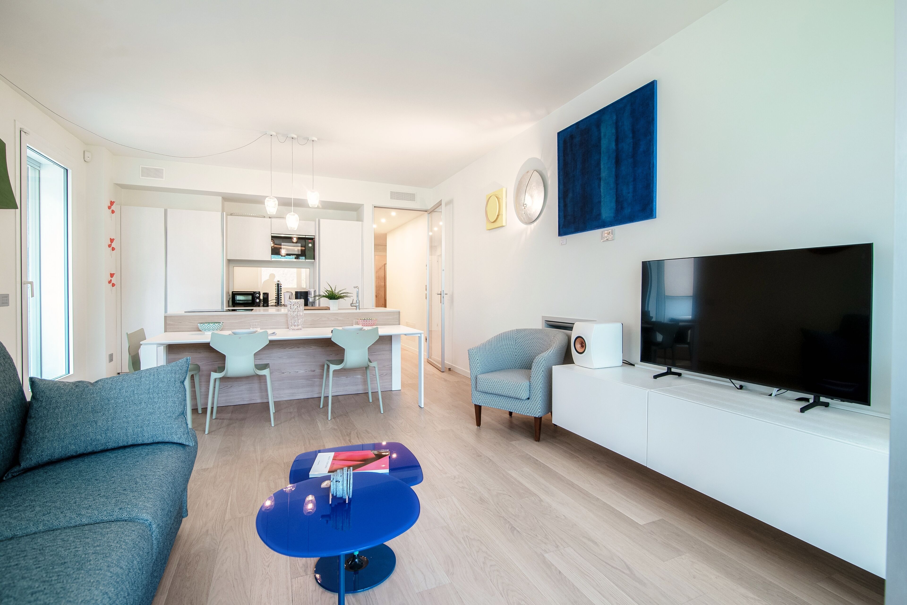 A modern living room with a sofa, a coffee table, a TV, and a dining area with chairs and a kitchen in the background.