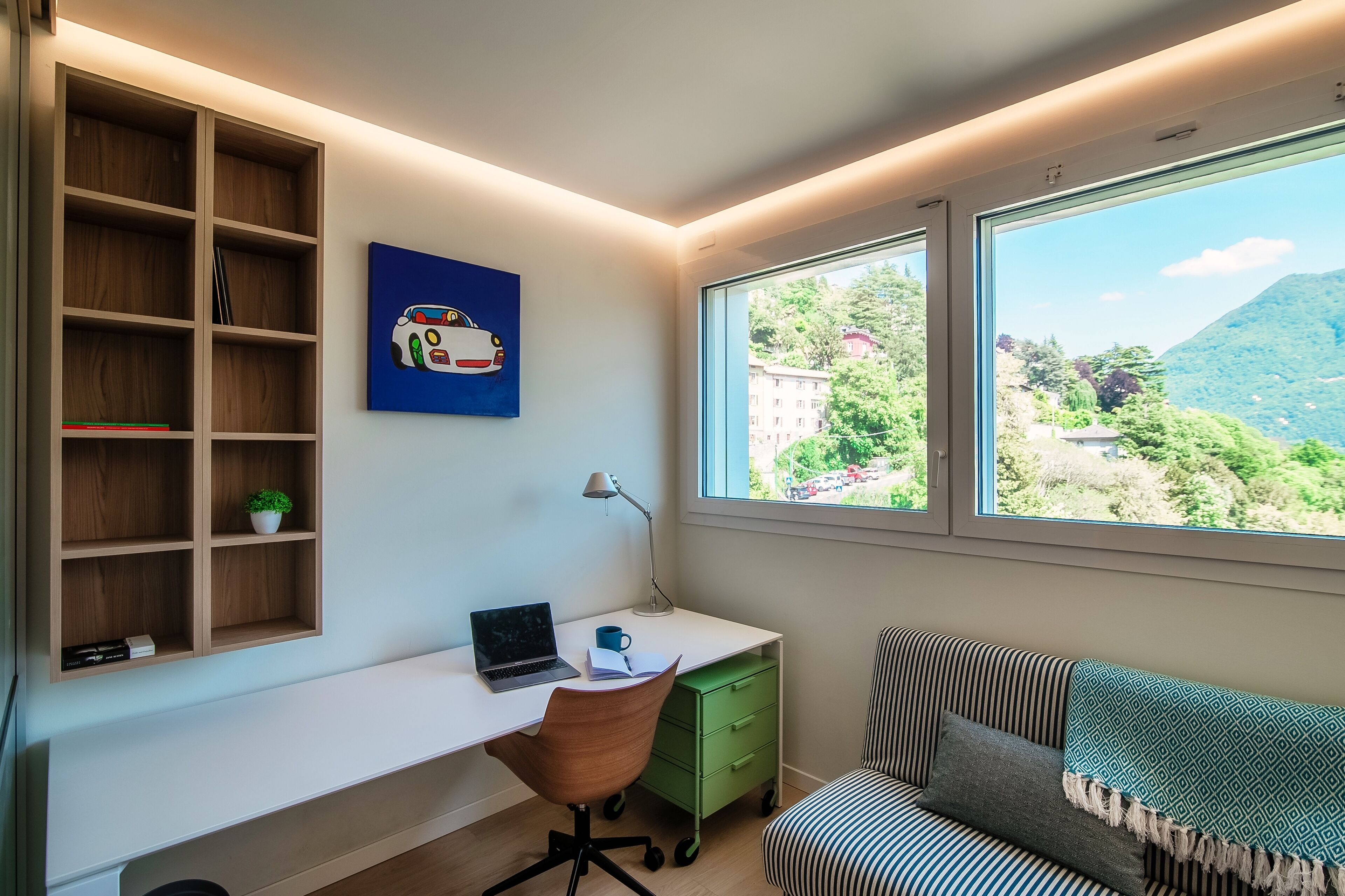 A modern room with a white desk, a laptop, a green drawer unit, a striped sofa, and a large window with a view of greenery and mountains.