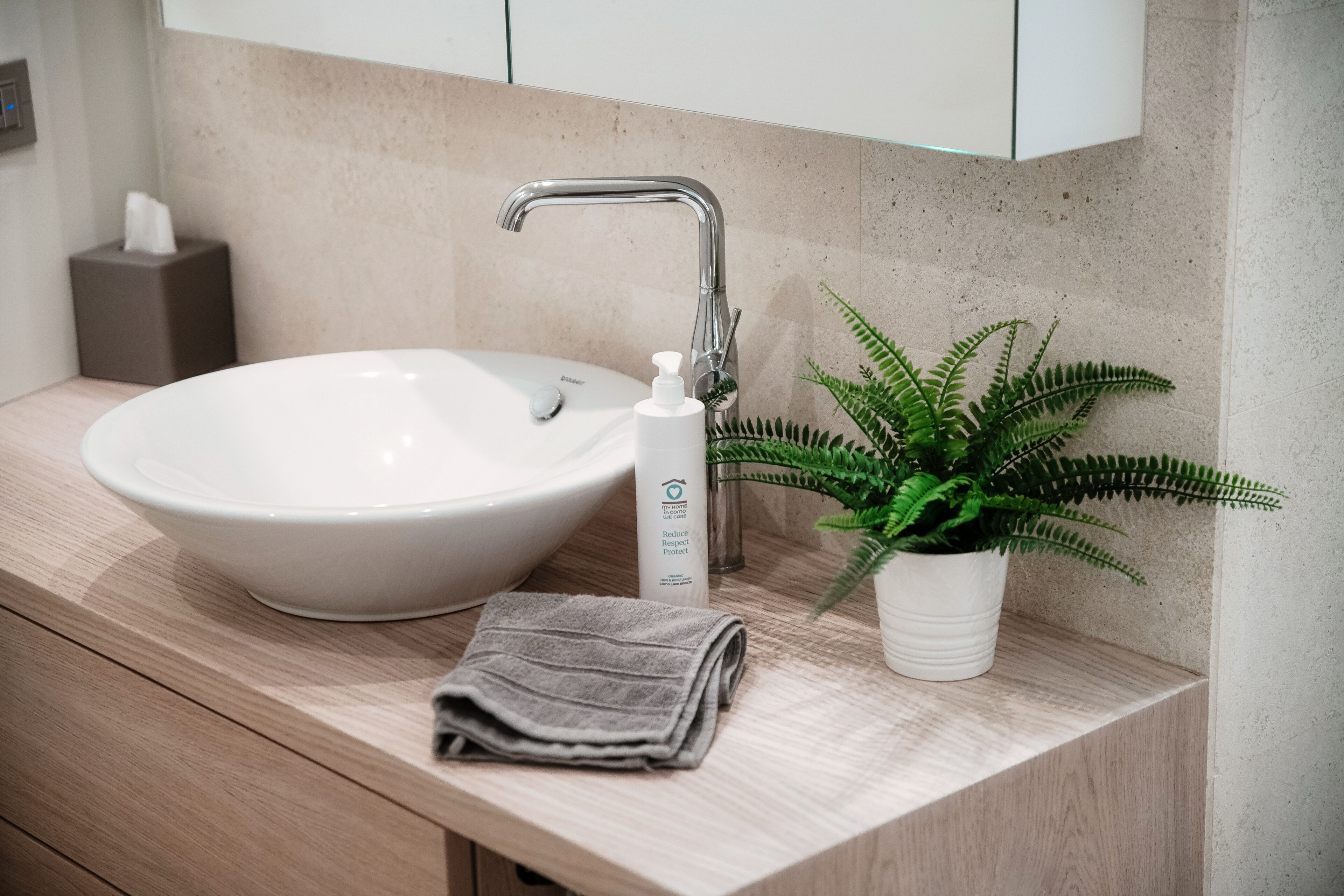Une salle de bain moderne avec un lavabo blanc, une serviette, un flacon de savon liquide et une fougère en pot.