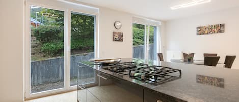 A modern kitchen with a central island, a dining area with chairs, and a view through large windows.