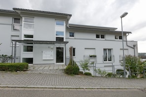 A modern two-story residential building with a flat roof, a front entrance with a glass door, and a small garden area with a metal fence.
