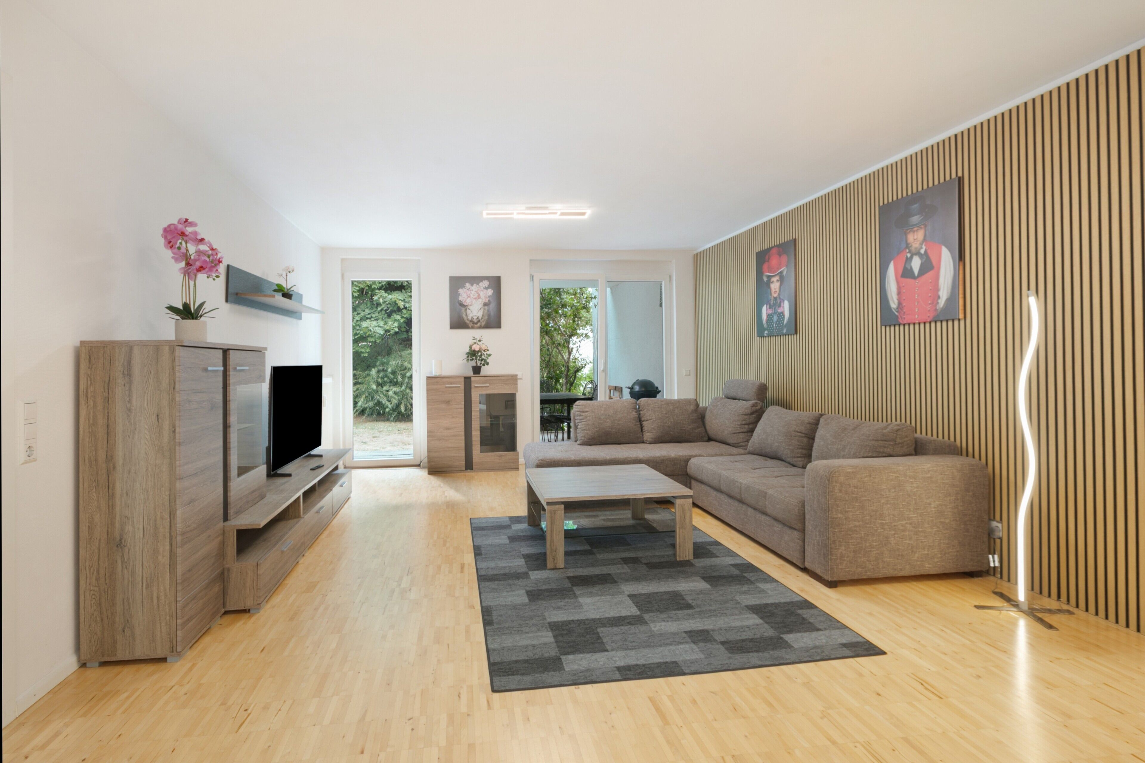 Ein modernes Wohnzimmer mit Flachbildfernseher, Holzmöbeln, einem grauen Teppich und einem großen Fenster.