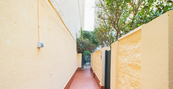 A narrow corridor with a red floor, flanked by yellow walls, leading to a green area with trees and a black gate.