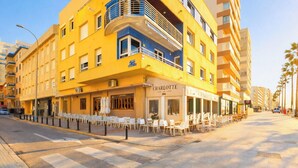 A yellow building with a café named 'Charlotte' on the ground floor, outdoor seating, and a pedestrian street with palm trees. - Double room - IMAR Hostel by Cadiz4Rentals (Cádiz)