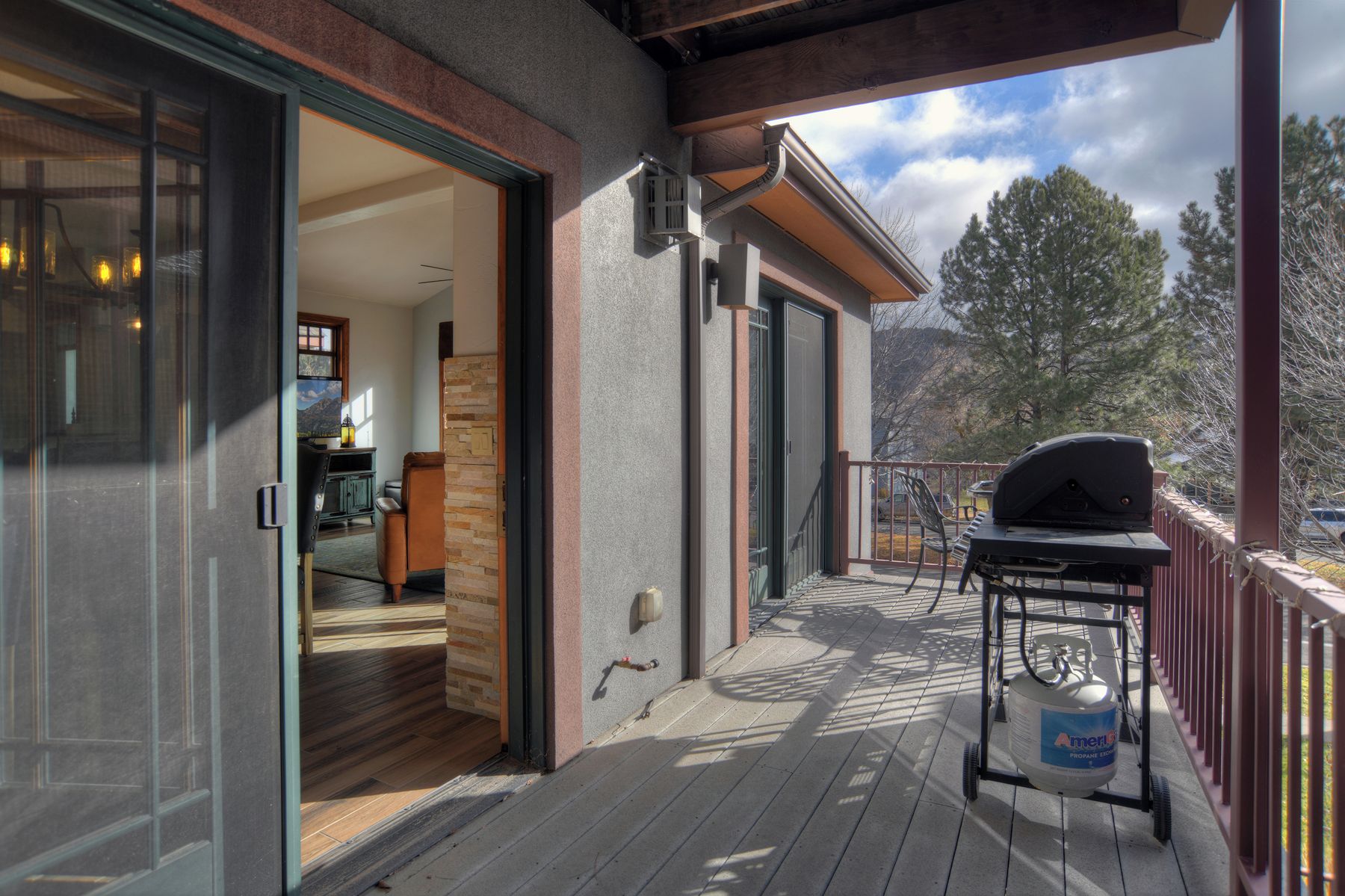 A covered deck with a barbecue grill and propane tank, a sliding glass door leading to an interior with a kitchen and seating area.