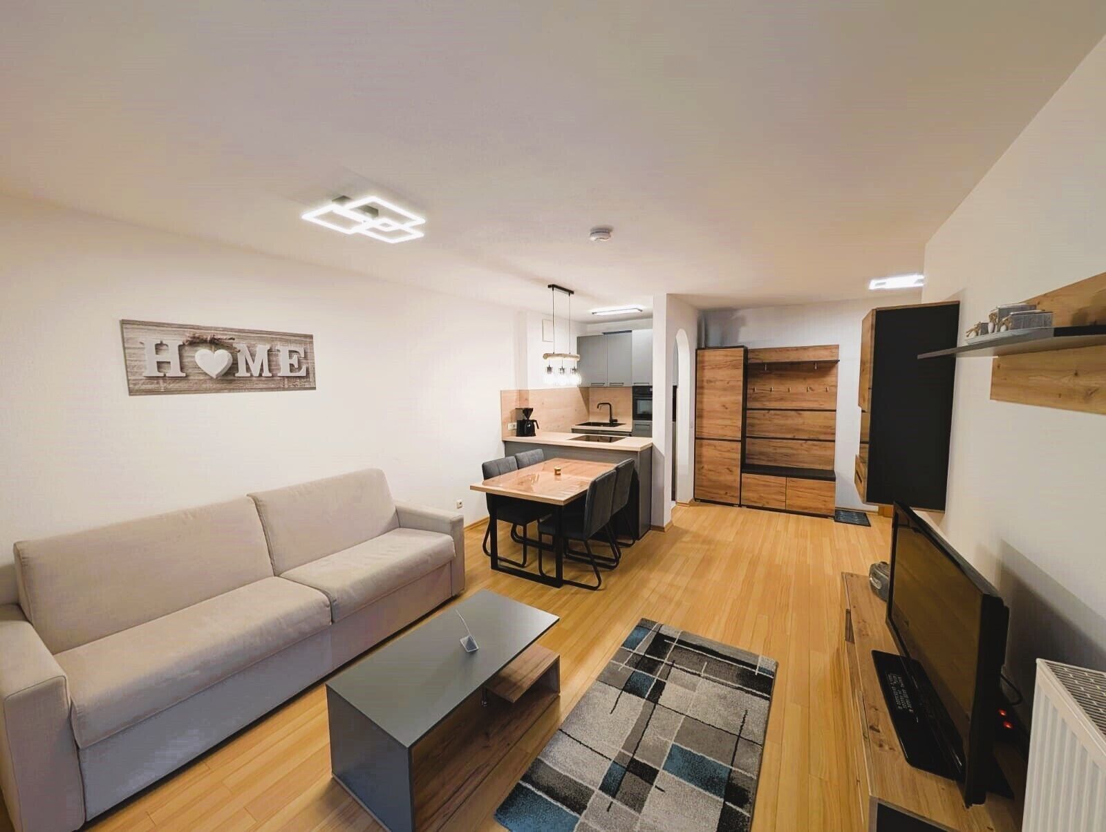 A modern living room with a sofa, coffee table, and a TV. There is a kitchen area with a dining table and chairs.