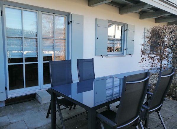 Une terrasse avec une table et des chaises, une fenêtre avec des volets et, au loin, la vue d’un bâtiment.