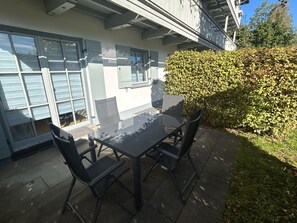 Une terrasse extérieure avec une table à plateau de verre et quatre chaises, entourée d’une haie et d’un bâtiment doté de grandes fenêtres.