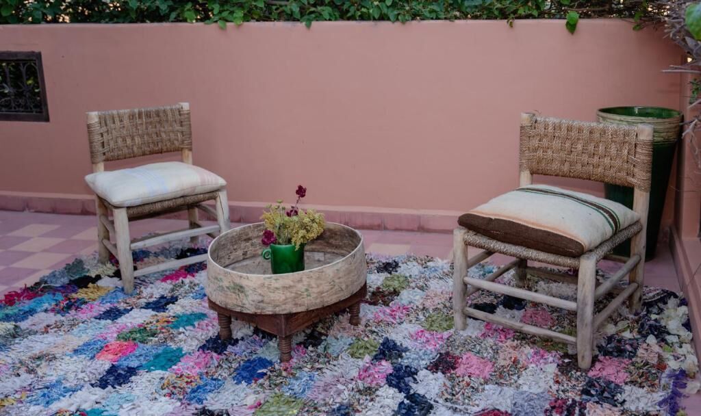 Two wicker chairs with cushions, a round wooden table with a floral arrangement, and a colorful, multicolored rug on a pink wall with greenery above.