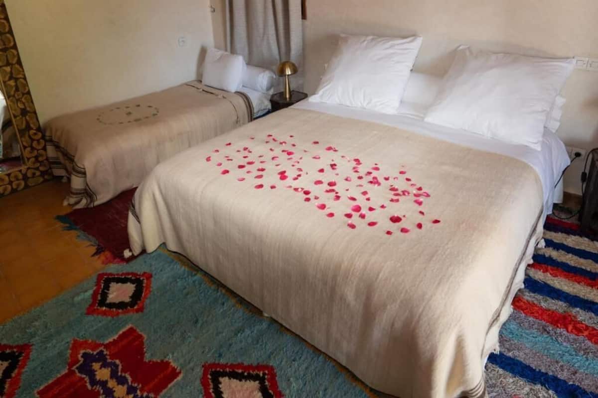 Ein Hotelzimmer mit zwei Betten, einem großen Bett mit weißer Bettdecke und rosa Rosenblättern sowie einem kleineren Bett mit gemusterter Bettdecke.