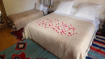 A hotel room with two beds, a large bed with a white bedspread and pink rose petals, and a smaller bed with a patterned bedspread.