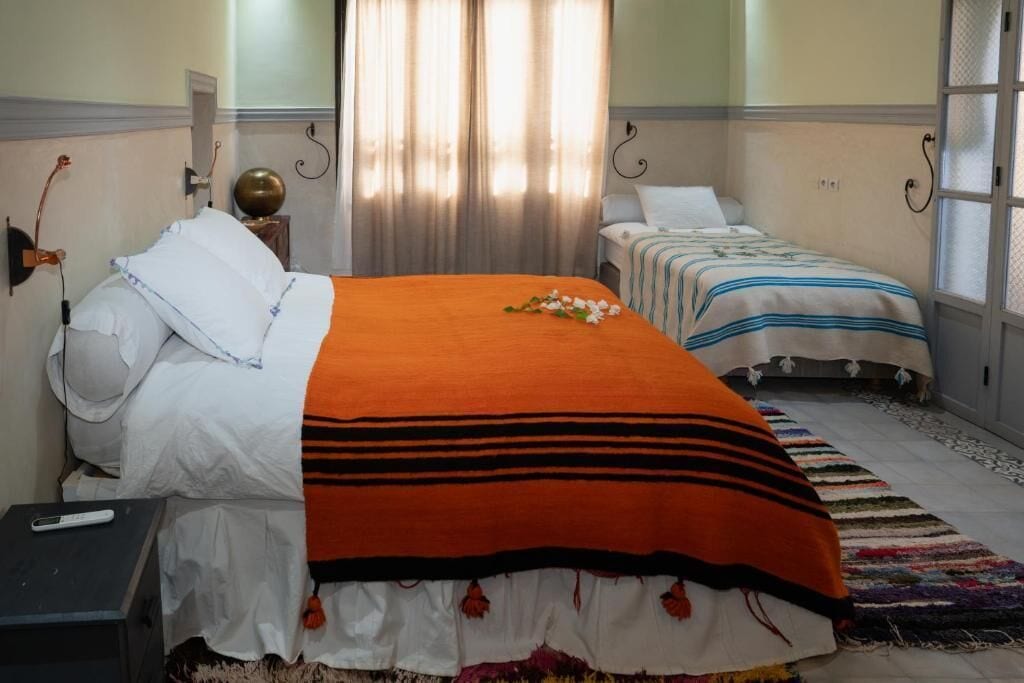 A hotel room with two beds, one with an orange blanket and the other with a striped blanket. There is a small table with a remote control on it, and a colorful rug on the floor.