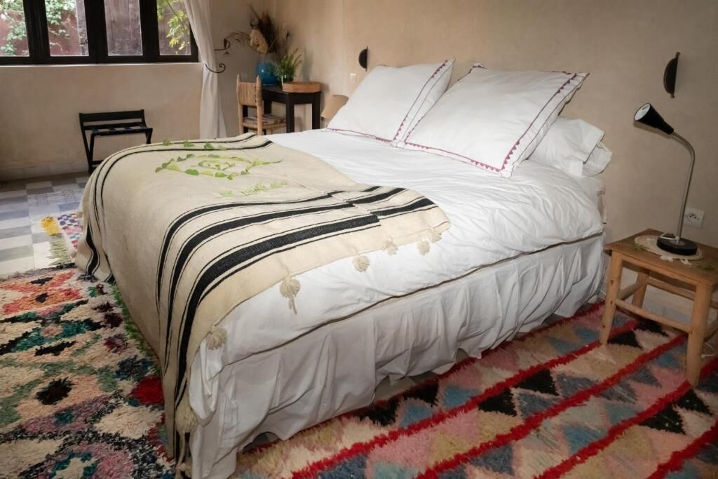 A bed with white linens and striped bedding, a small bedside table, a lamp, and a colorful rug.