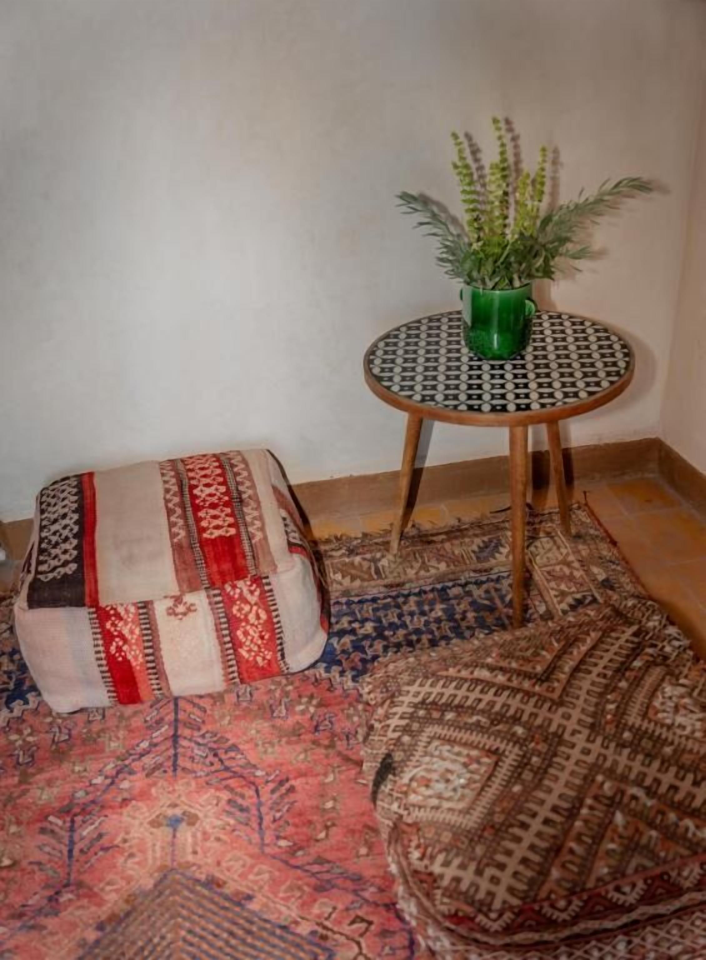 A room with a patterned rug, a small round table with a green plant, and a traditional woven pouf.