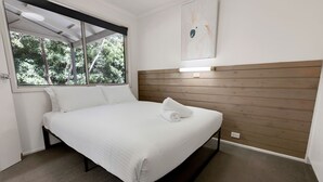 Un lit bien fait, avec du linge de lit blanc, une tête de lit en bois et une fenêtre donnant sur des arbres.