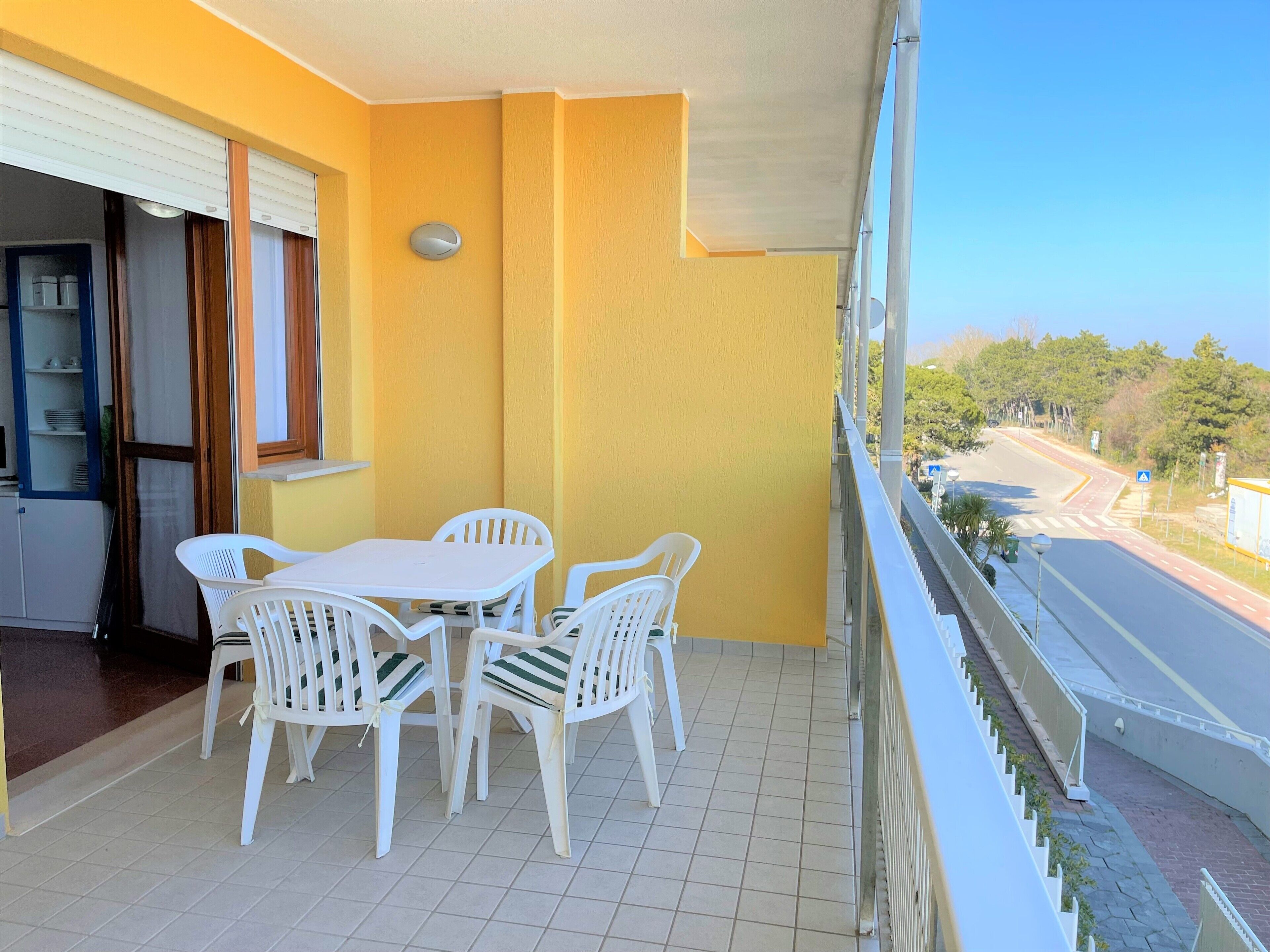 A balcony with a yellow wall, a white table with chairs, and a view of a road and greenery.
