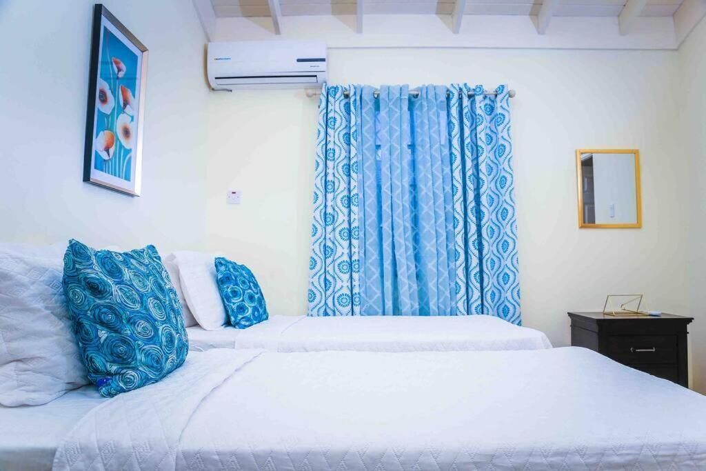 A bedroom with a bed, pillows, curtains, a wall-mounted air conditioner, a framed picture, and a small bedside table.
