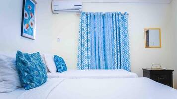 A bedroom with a bed, pillows, curtains, a wall-mounted air conditioner, a framed picture, and a small bedside table.