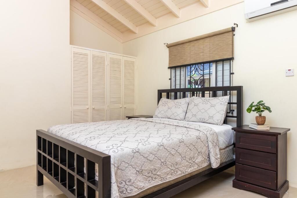 Une chambre à coucher avec un lit, une table de chevet, une plante en pot, une fenêtre avec des stores et une armoire blanche.