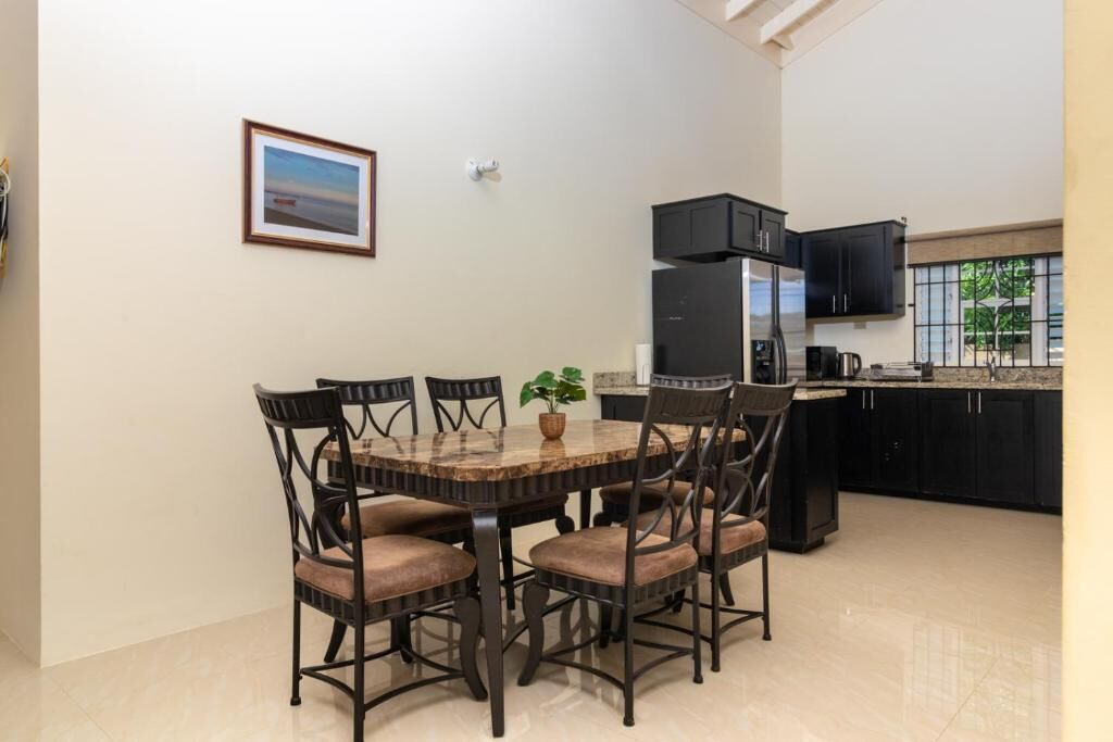 Un comedor con una mesa redonda, seis sillas, una planta en maceta, y una cocina con armarios negros y un refrigerador.
