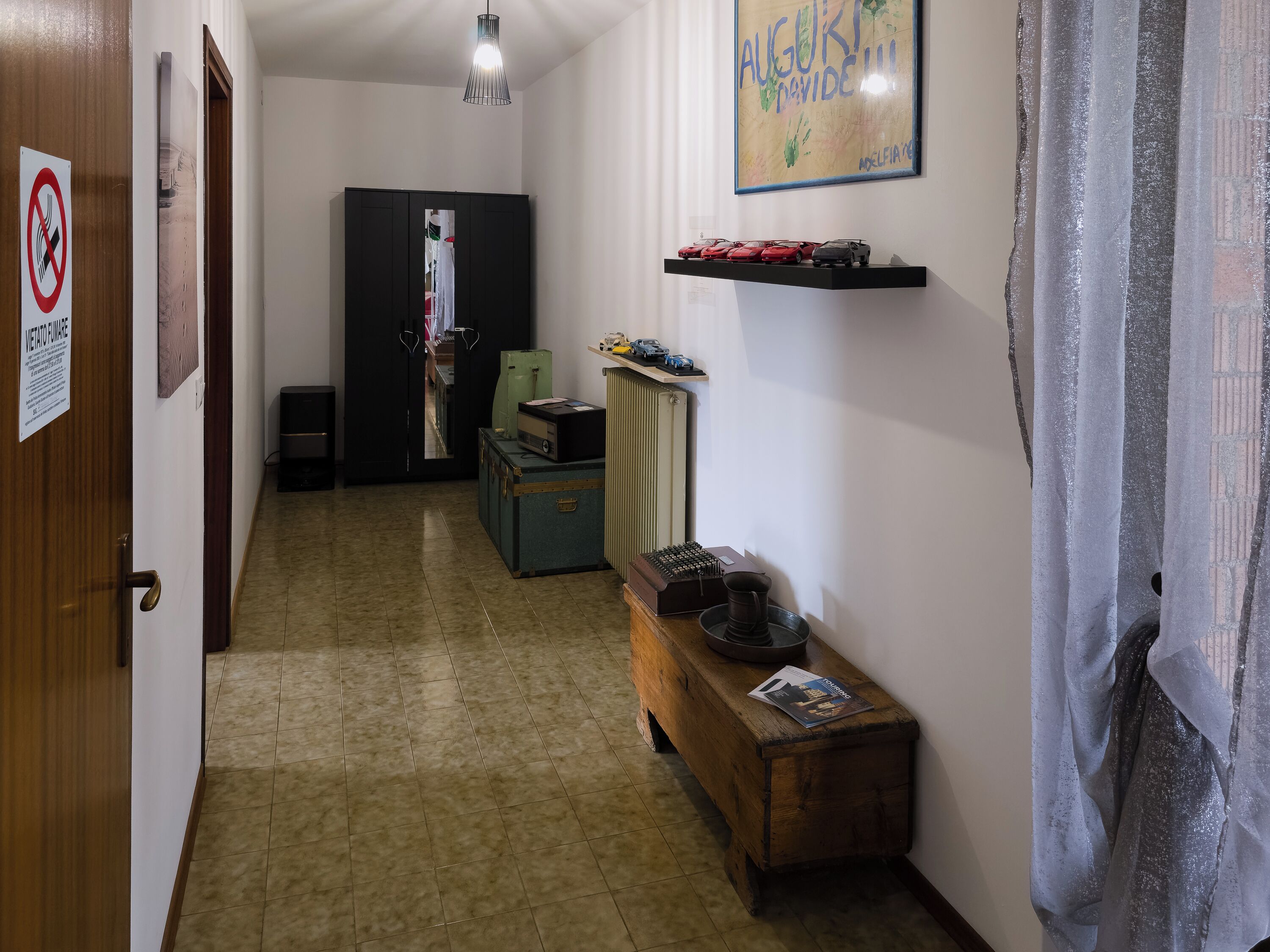 Un pasillo con una puerta de madera, un cartel de prohibido fumar, un armario negro, un escritorio con una computadora y una estantería con maquetas de coches.