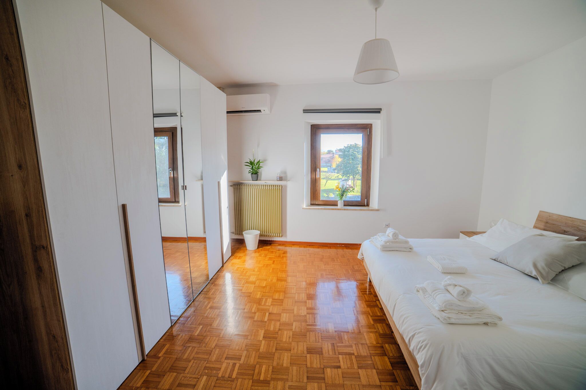 A bedroom with a wooden floor, a bed with white linens, a radiator, a potted plant, and a window with a view of greenery.