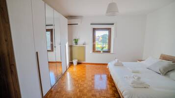 A bedroom with a wooden floor, a bed with white linens, a radiator, a potted plant, and a window with a view of greenery.