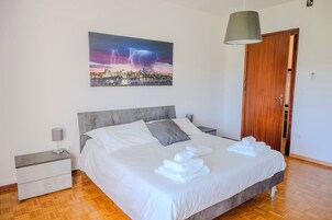 Una cama bien hecha con sábanas y toallas blancas, cabecera gris, mesita de noche con lámpara y un cuadro de paisaje urbano en la pared.
