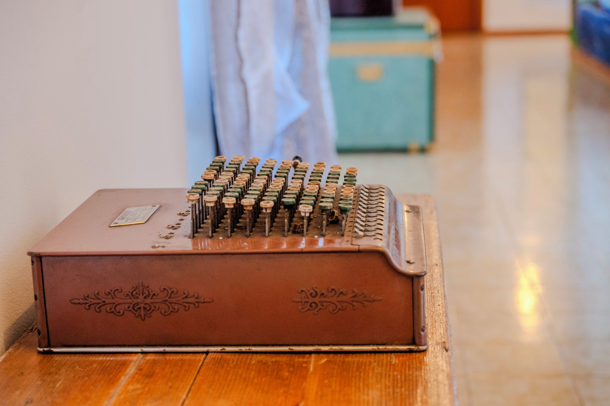Een oude schrijfmachine op een houten tafel in een kamer met een blauwe koffer en een witte gordijn in de achtergrond.