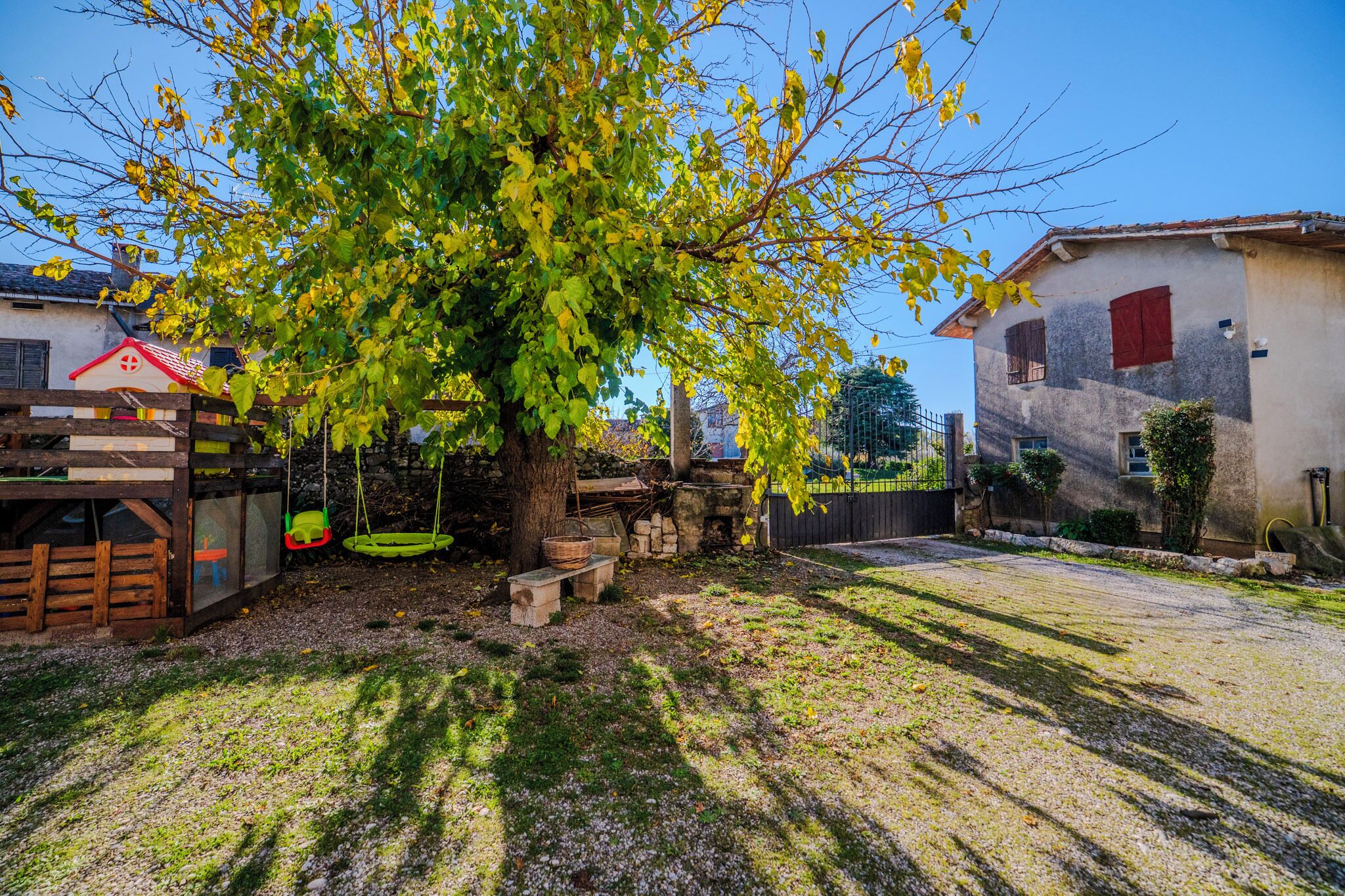 Un espacio exterior rústico con una casita de juegos de madera, un columpio y un gran árbol que da sombra.