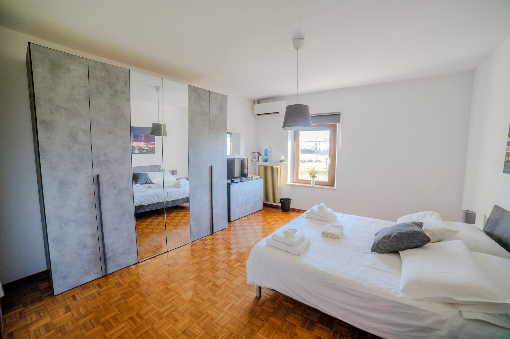 A modern bedroom with a large mirror, a bed with neatly folded towels, a dresser, and a window with a view.