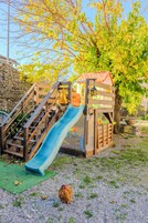 Una casita de juegos de madera con un tobogán, un muro de piedra y un árbol con hojas amarillas.