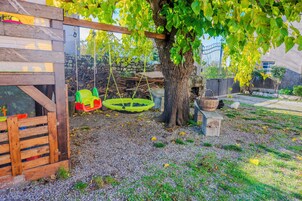 Una casita de madera con un columpio, un árbol y un jardín cercado.