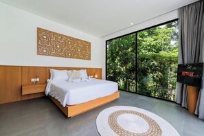 A modern bedroom with a large bed, a decorative wall panel, a view of greenery through a sliding door, and a Netflix sign. - Villa Talun by GORO (Kabupaten Gianyar)