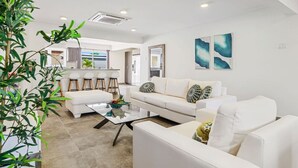 Een moderne woonkamer met een witte bank, een glazen salontafel en een keuken op de achtergrond.