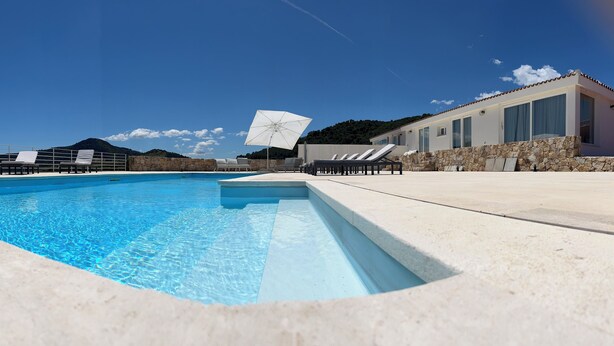 Una piscina con sillas de descanso y sombrillas, un muro de piedra y un edificio con ventanas grandes.