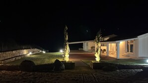 Vista nocturna de una propiedad costera con muro de piedra, dos árboles iluminados y un edificio blanco con porche cubierto.