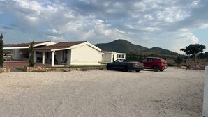 Una casa de dos pisos con un patio, un coche rojo y una montaña al fondo.