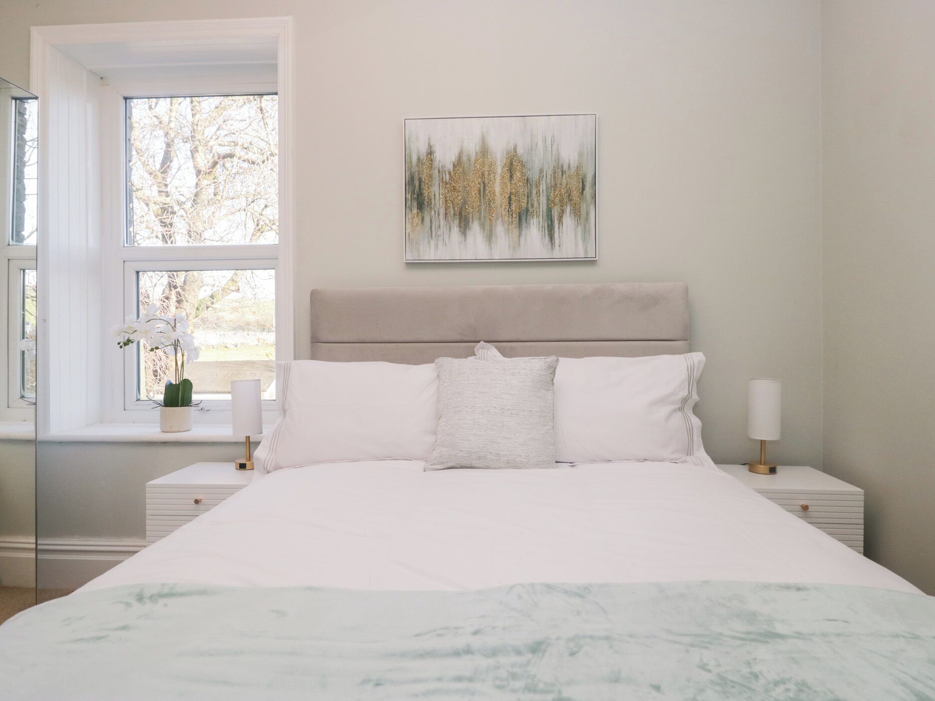 Une chambre à coucher avec un lit, une fenêtre donnant sur des arbres, une table de chevet avec une lampe et un tableau accroché au mur.